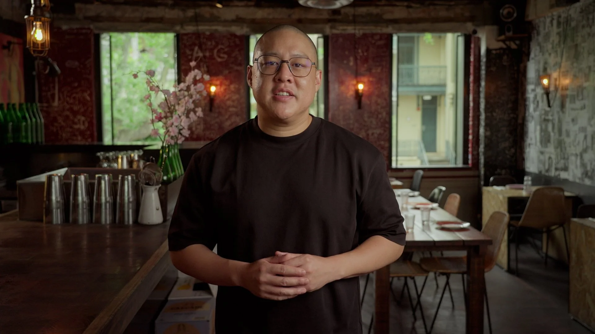 A man with glasses and a black shirt standing inside a restaurant with a brick wall, large windows, and dining tables in the background.