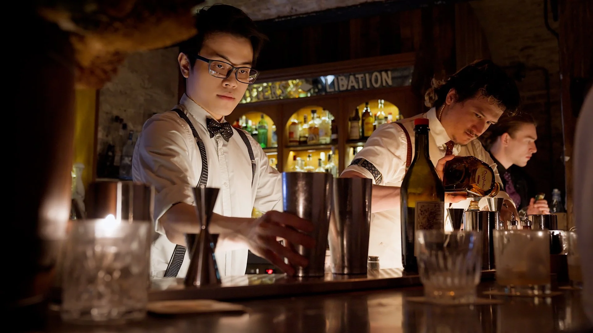 A bartender and two baristas working behind a bar counter in a dimly lit bar or cocktail lounge, preparing drinks with various bottles and bar tools.