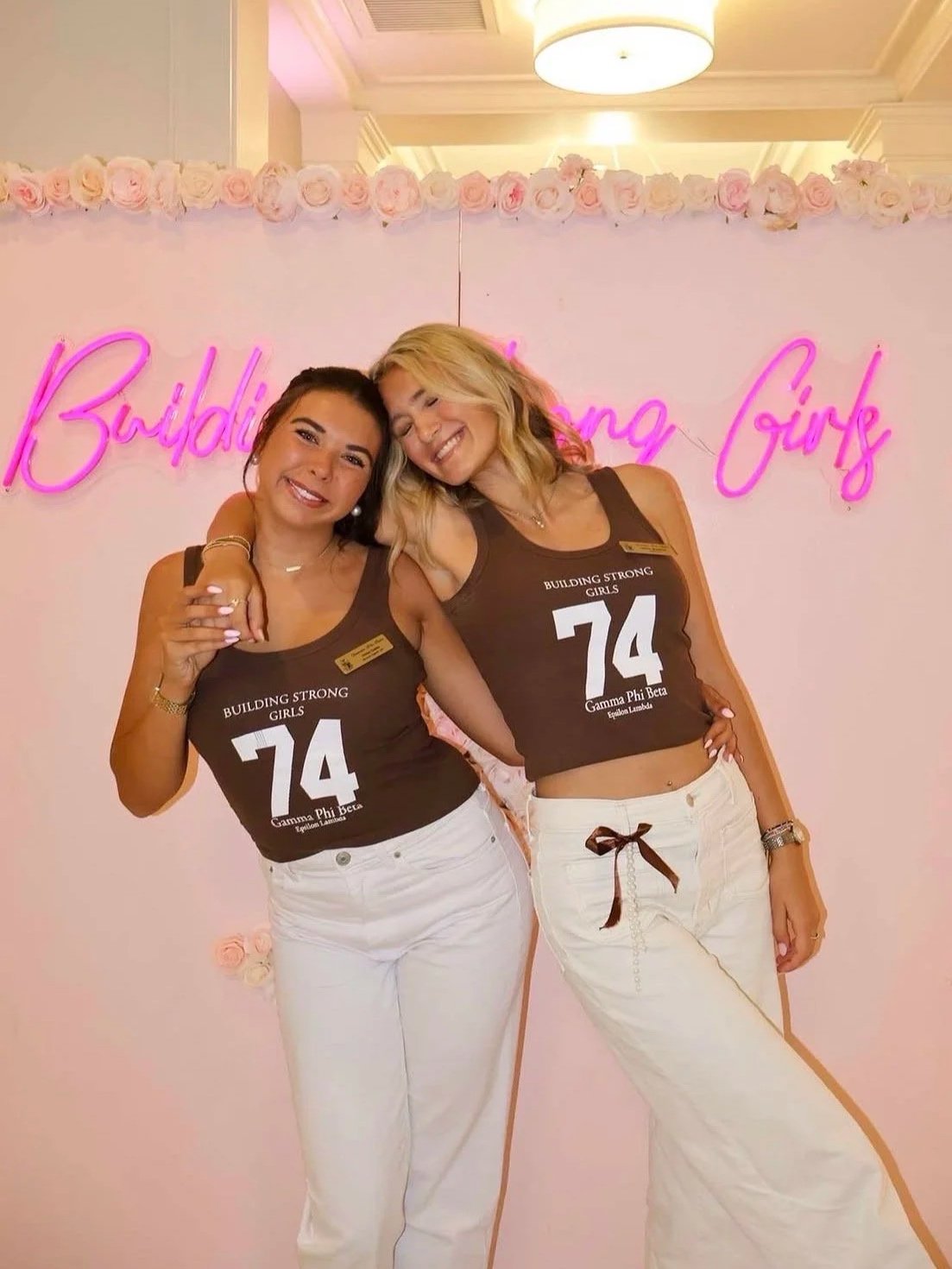 Two women in brown tank tops with white pants standing in front of a pink wall with a floral border and neon sign that reads 'Building Strong Girls'. They are smiling and hugging, one with her arm around the other's shoulder.