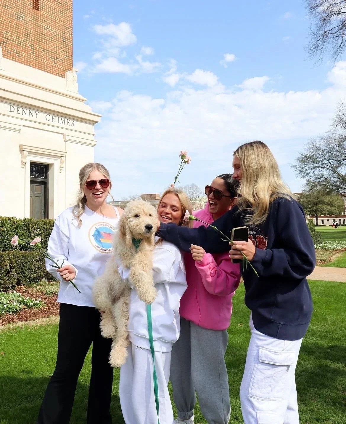 Four young women and a dog outdoors on a grassy area, smiling and holding flowers, in front of a building with a sign that reads 'Denny Chimes.' The women are dressed casually, and the dog appears to be a large, fluffy, light-colored breed, sitting i