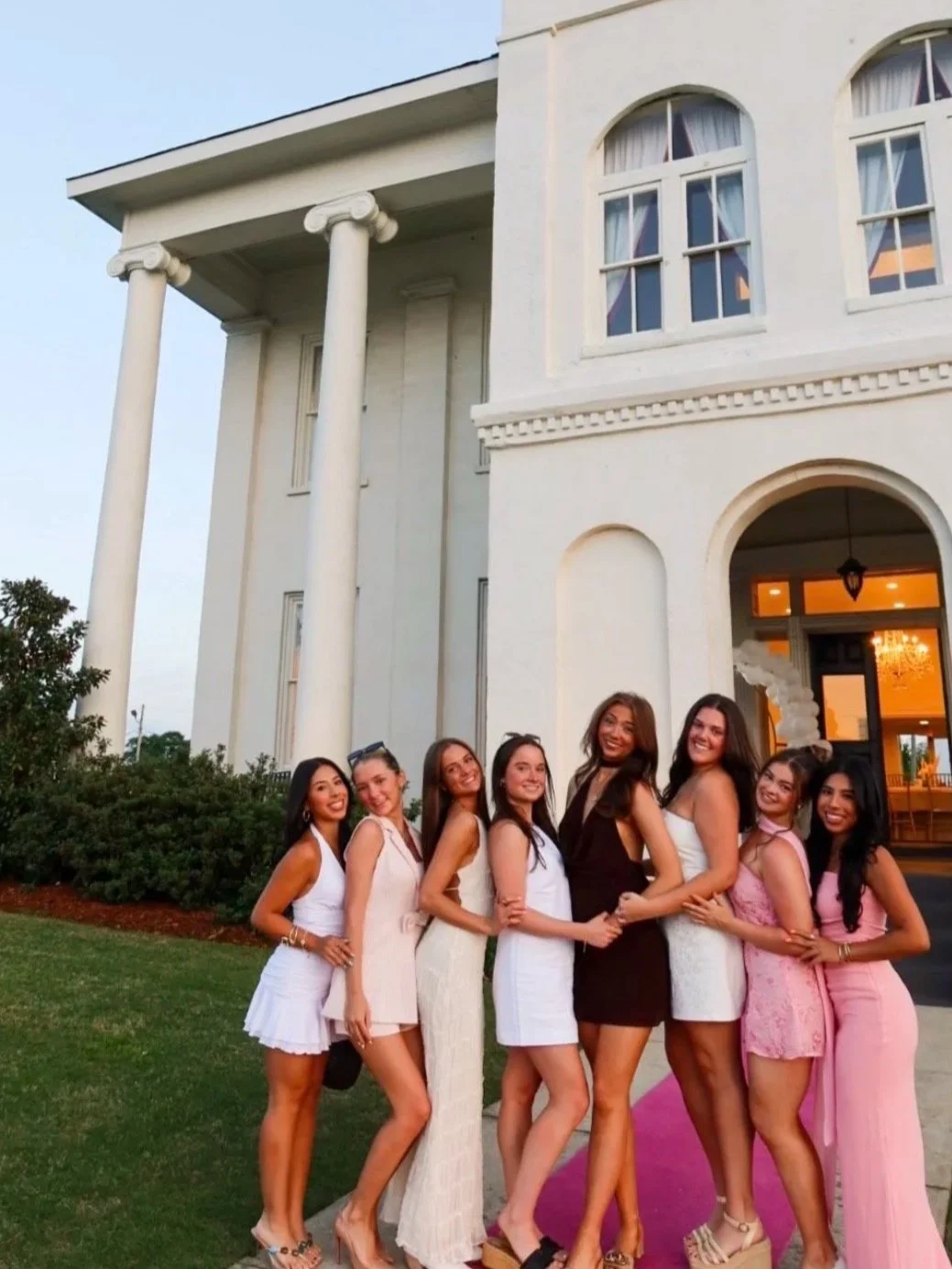 Group of women standing on a pink carpet outside a white mansion with large windows and columns during sunset.