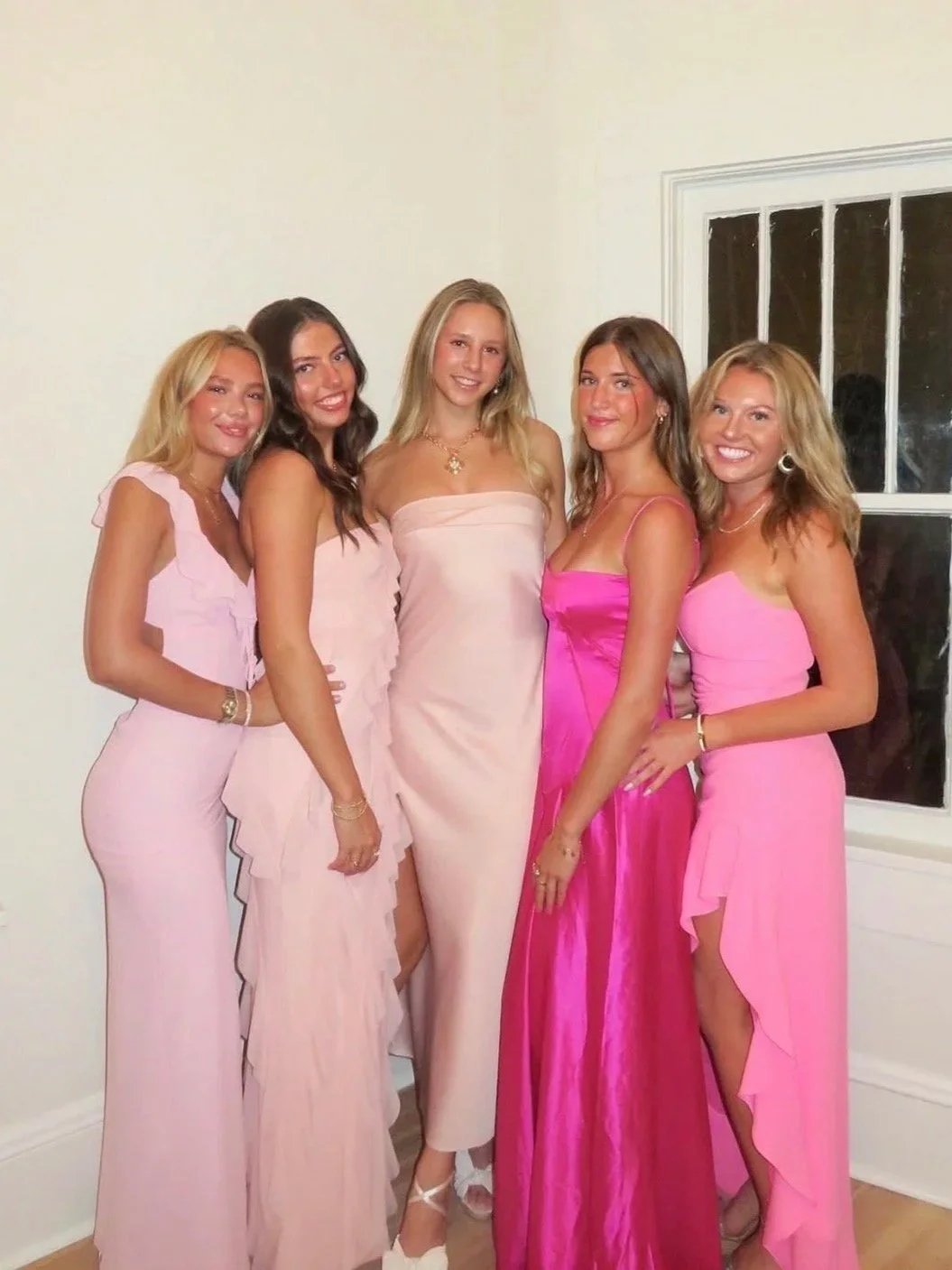 Five young women wearing pink and peach formal dresses at a social gathering, standing close together inside a room with white walls and a window in the background.