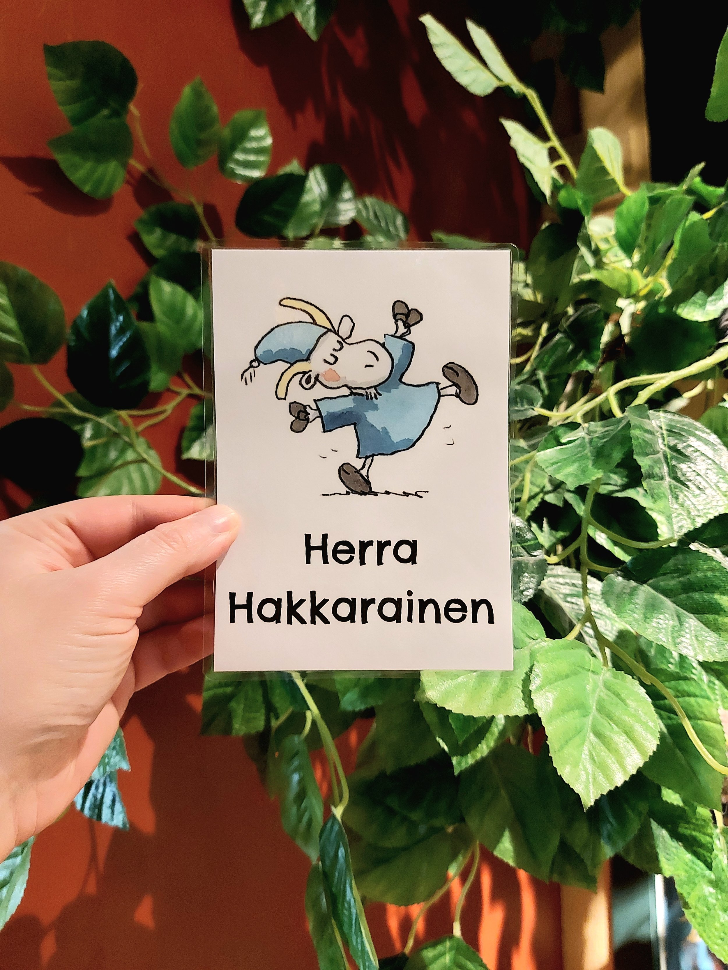 Valokuva paperikortista, jossa on vuohihahmo Herra Hakkaraisen kuva ja nimi. Taustalla sisätiloissa oleva tekopuu.