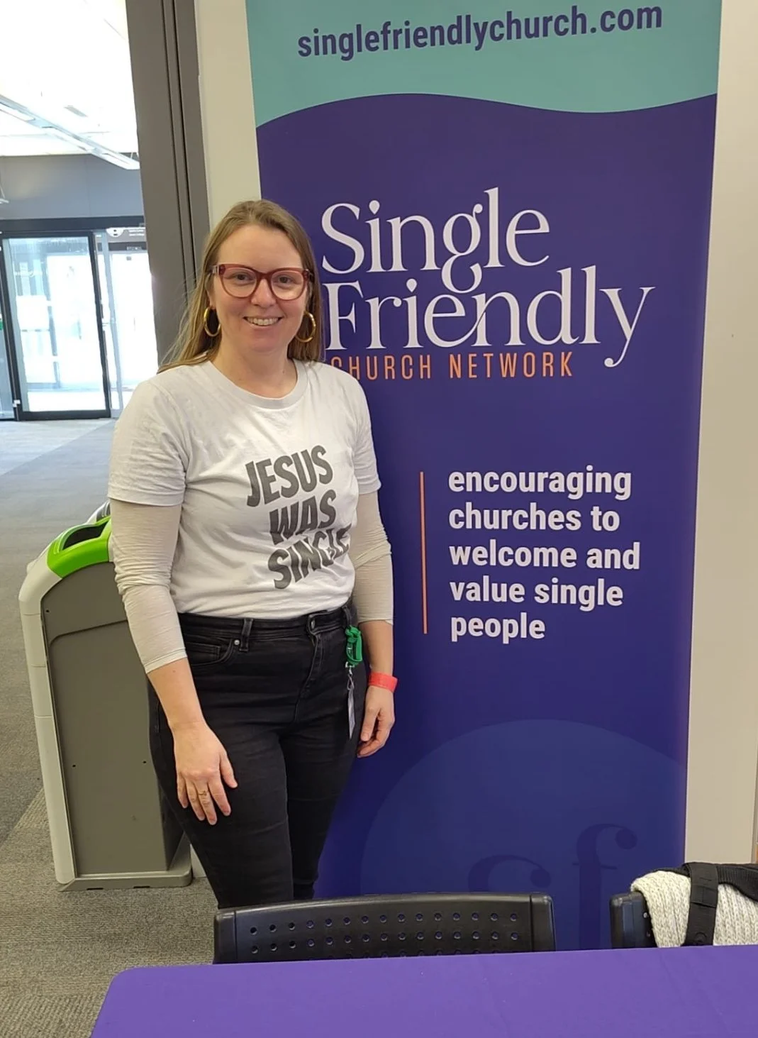 Liz Dunbar at Flourish Festival, standing by the Single Friendly Church Network banner. She is wearing a t-shirt with 'Jesus was Single' writing