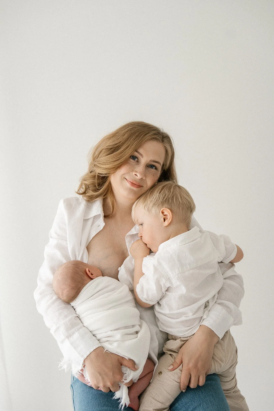 Äiti imettää vastasyntynyttä ja on syliin ottanut myös kaksi pienenpienempää lasta, yksi istuu hänen sylissään ja toinen istuu hänen polvellaan. Perhe hymyilee ja katsoo kameraan.