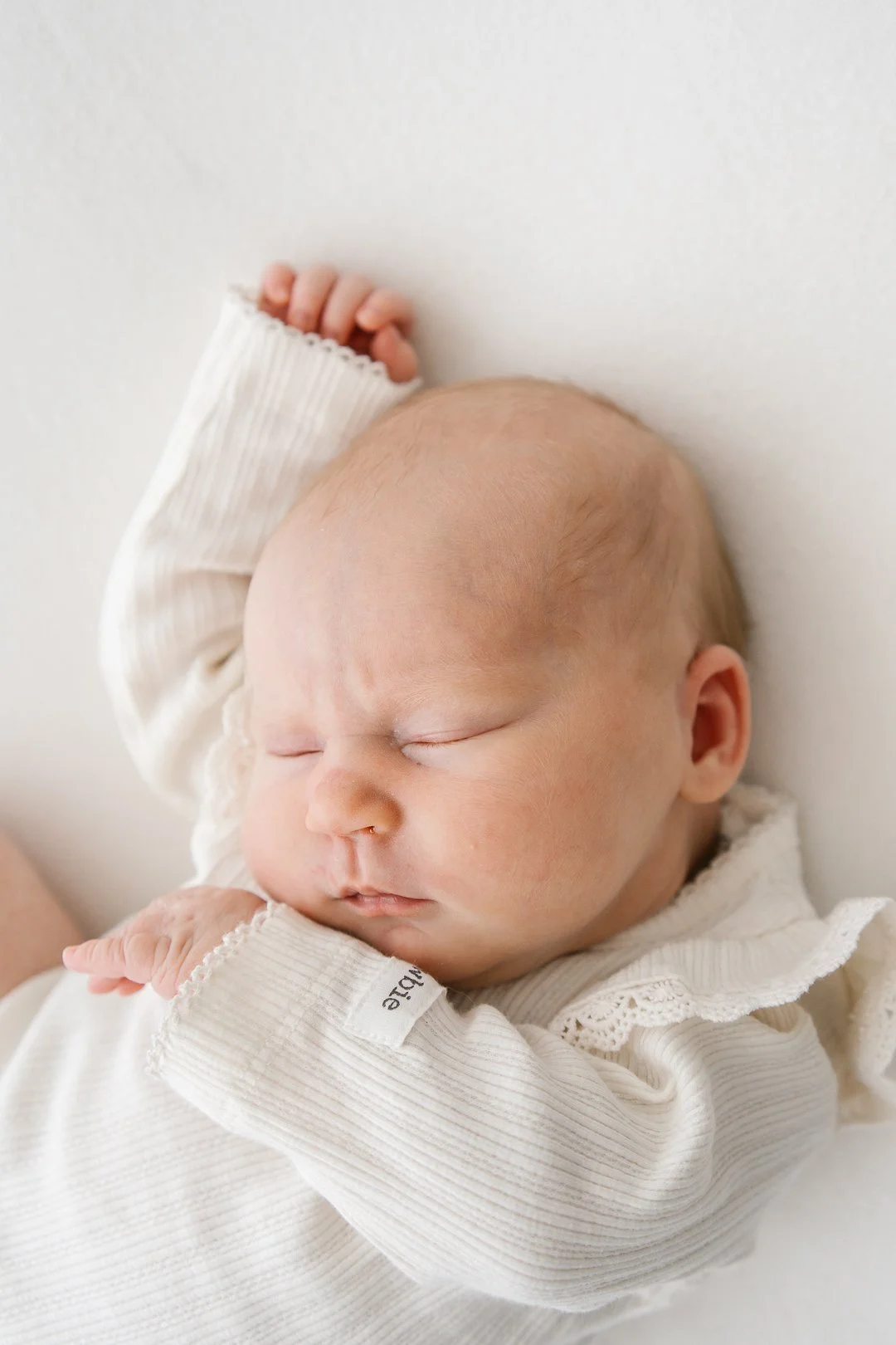 Vauva nukkuu, makaa valkoisella taustalla, yllään valkoinen pitkähihaisen puseron. Vauvan kasvot ovat rauhalliset ja silmät kiinni.