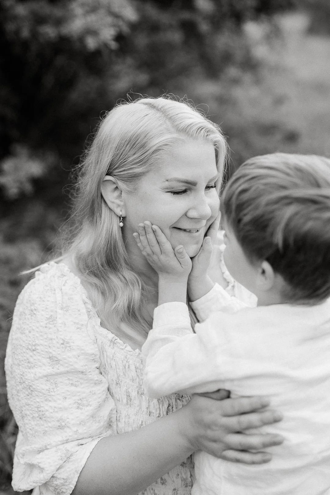 Nainen ja lapsi lähekkäin, nainen hymyilee ja lapsen kädet voivat olla kasvoilla, kuvassa on mustavalkoisen kuva.