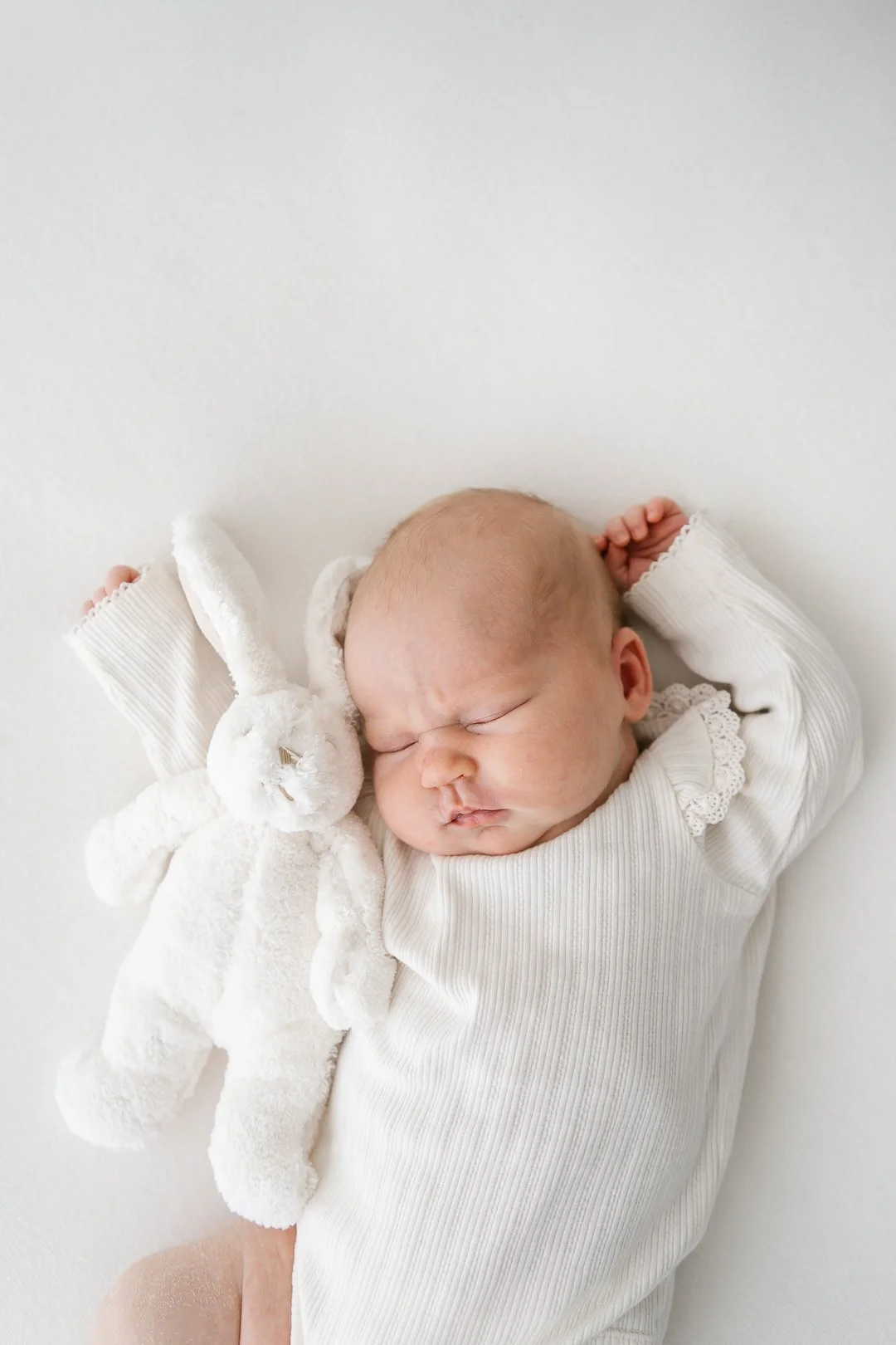 Pieni vastasyntynyt nukkumassa, vieressään pehmolelu, vaaleissa vaatteissa, valkoisella taustalla.