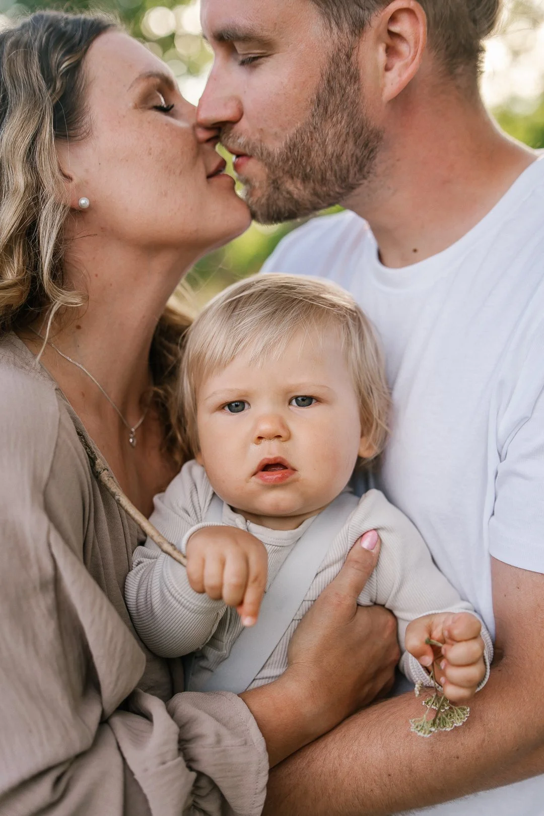 Perhe noin kahden aikuisen ja yhden lapsen kanssa ulkona, aikuiset suutelevat toisiaan, lapsi katsoo suoraan kameraan.