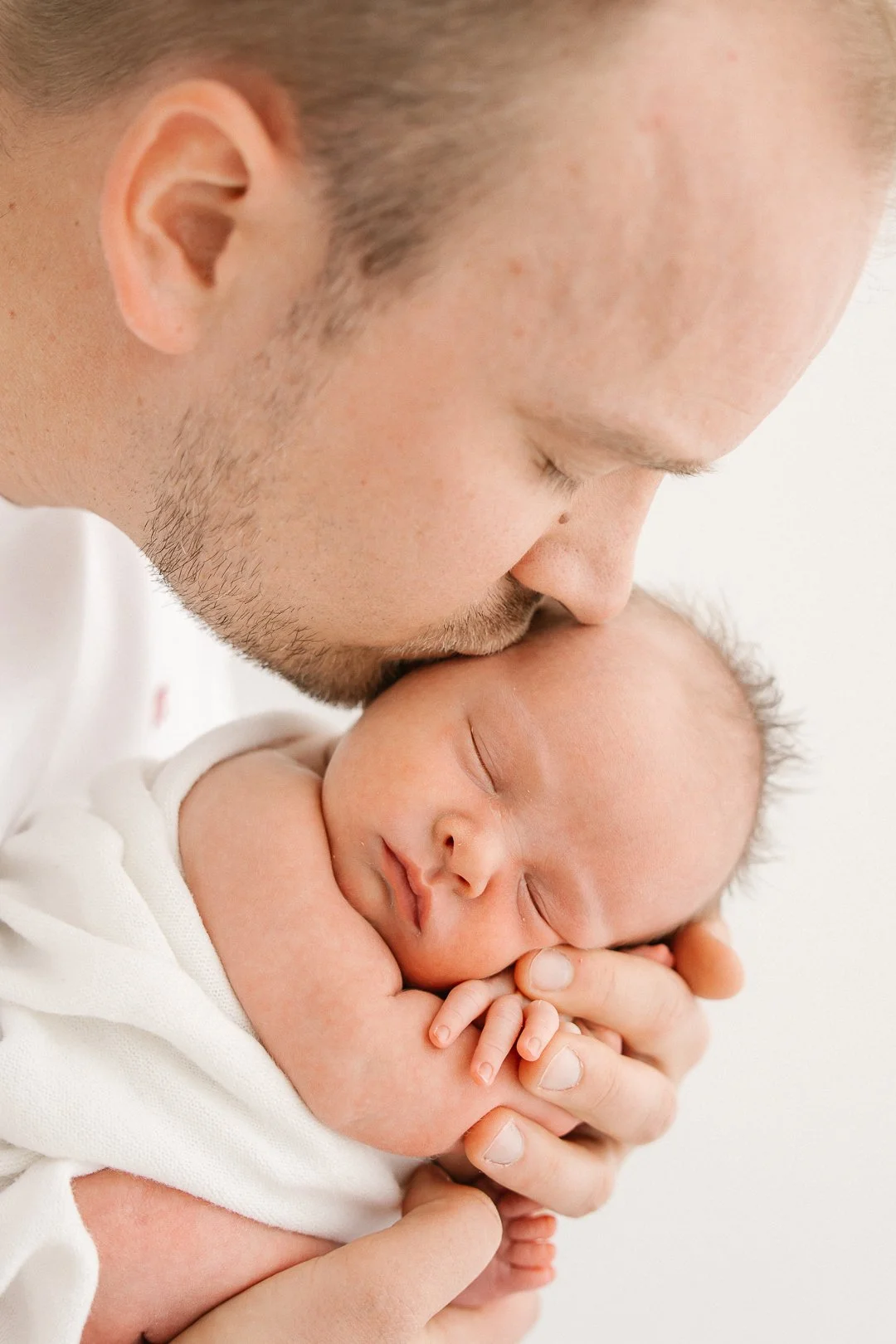 Vauva nukkuu käpertyneenä vanhemman käsiin, vanhempi suutelee lastaan otsalle.