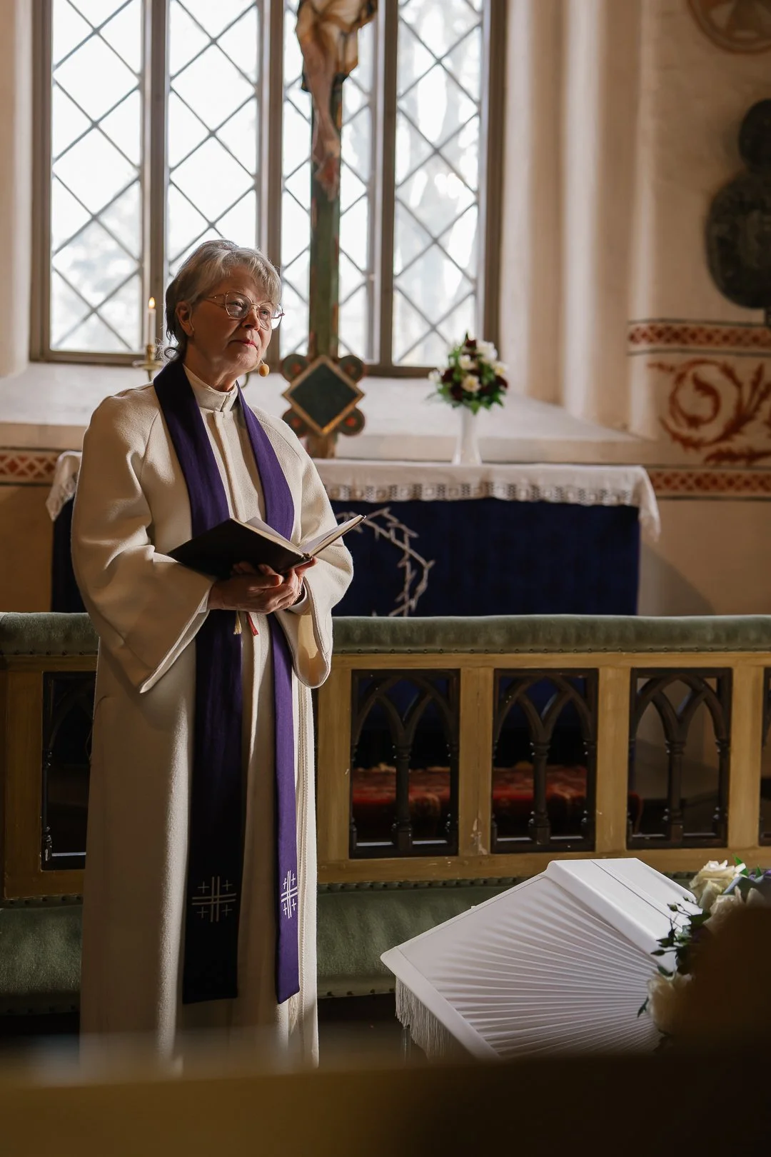 Nainen pappi seisoo kirkossa, lukee kirjaa, taustalla risti ja ikkunat.