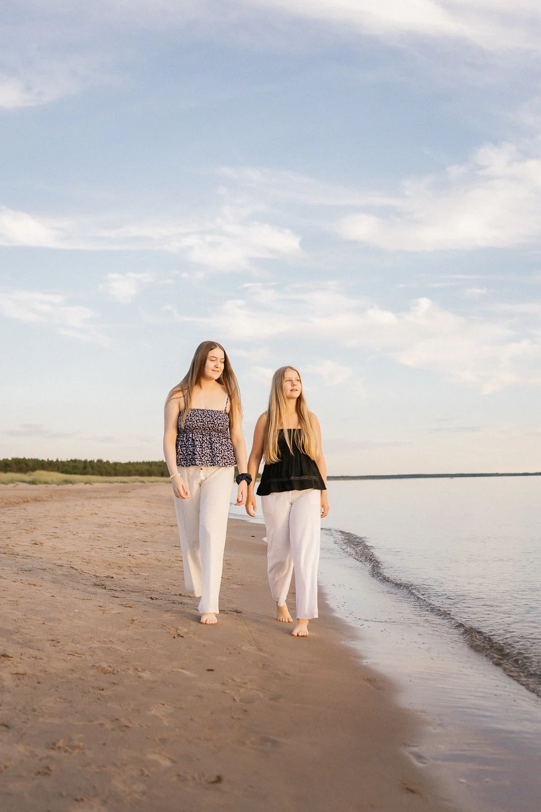 Kaksi nuorta naista kävelee rannalla meren äärellä, taivas on pilvinen ja meri on rauhallinen.