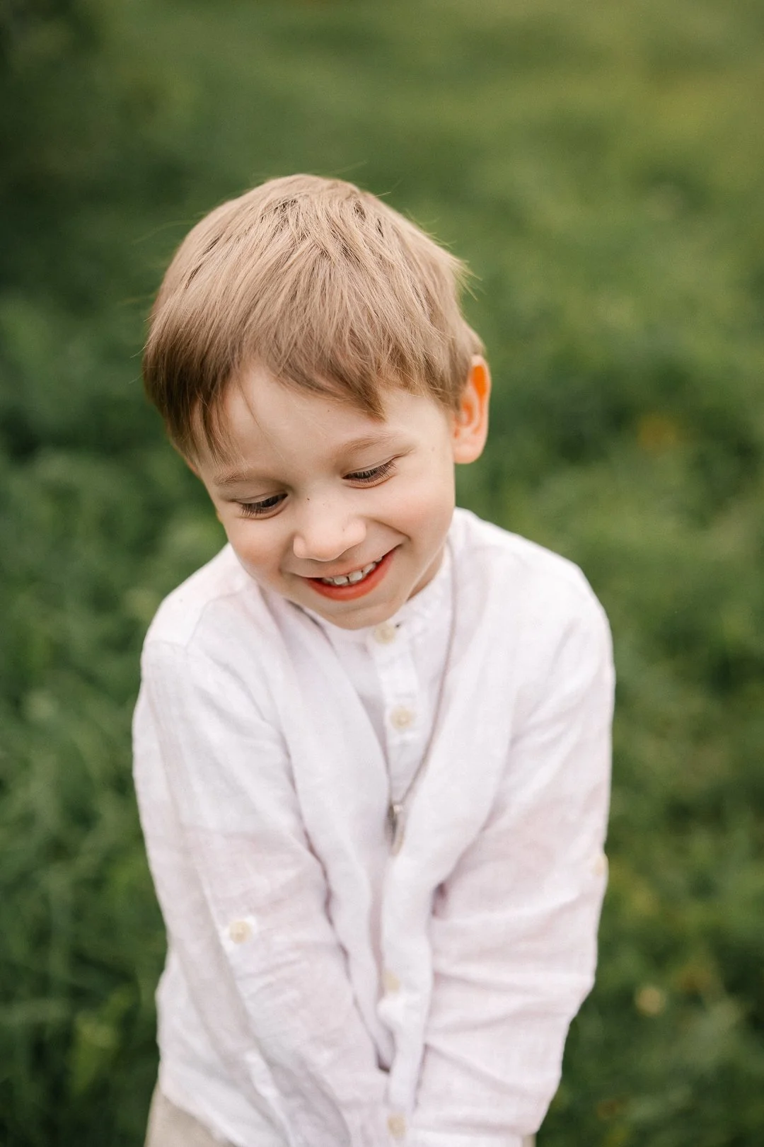 Hymyilevä poika, jolla on vaalea hiusty, vaaleahiuksinen, ja valkoinen pusero, seisoo vihreän taustan edessä ulkona.