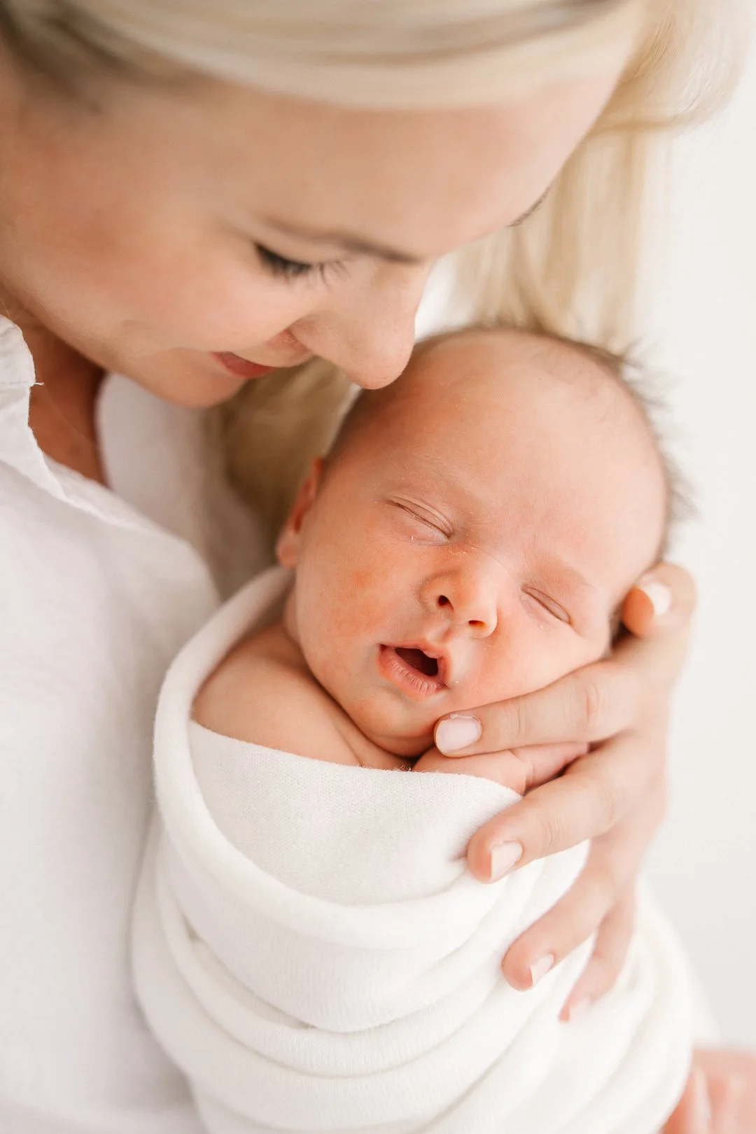 Nuori nainen ja vastasyntynyt vauva, nainen hymyilee lämpimästi ja fetus-asennossa oleva vauva on sylissä, kiedottuna valkoiseen peittoon.