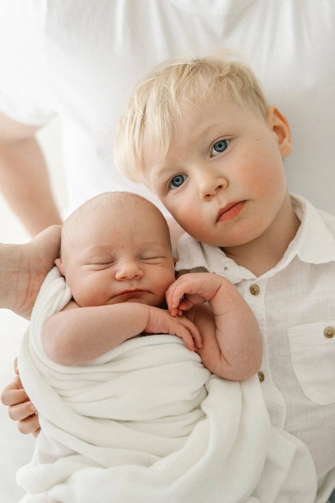 Nuori poika, jolla on vaaleat hiukset ja siniset silmät, pitää sylissään vastasyntynyttä vauvaa, joka on nukkuva ja kääritty valkoiseen peittoon.