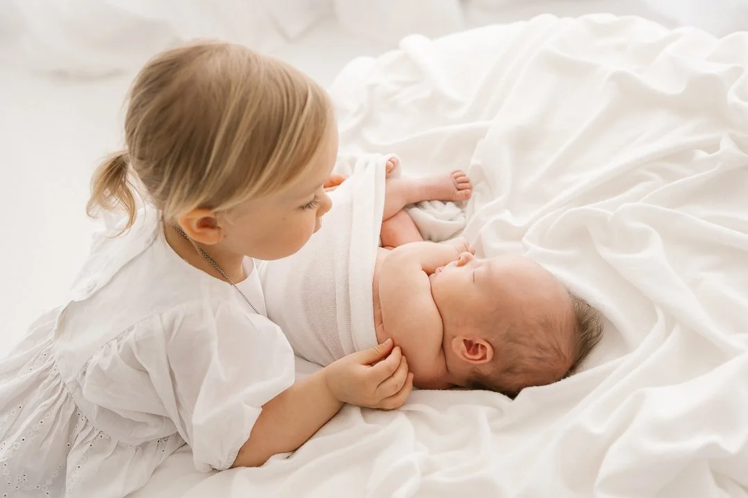 Pikkutyttö katsoo hellästi vastasyntynyttä siskoa, joka makaa valkoisella vuoteella