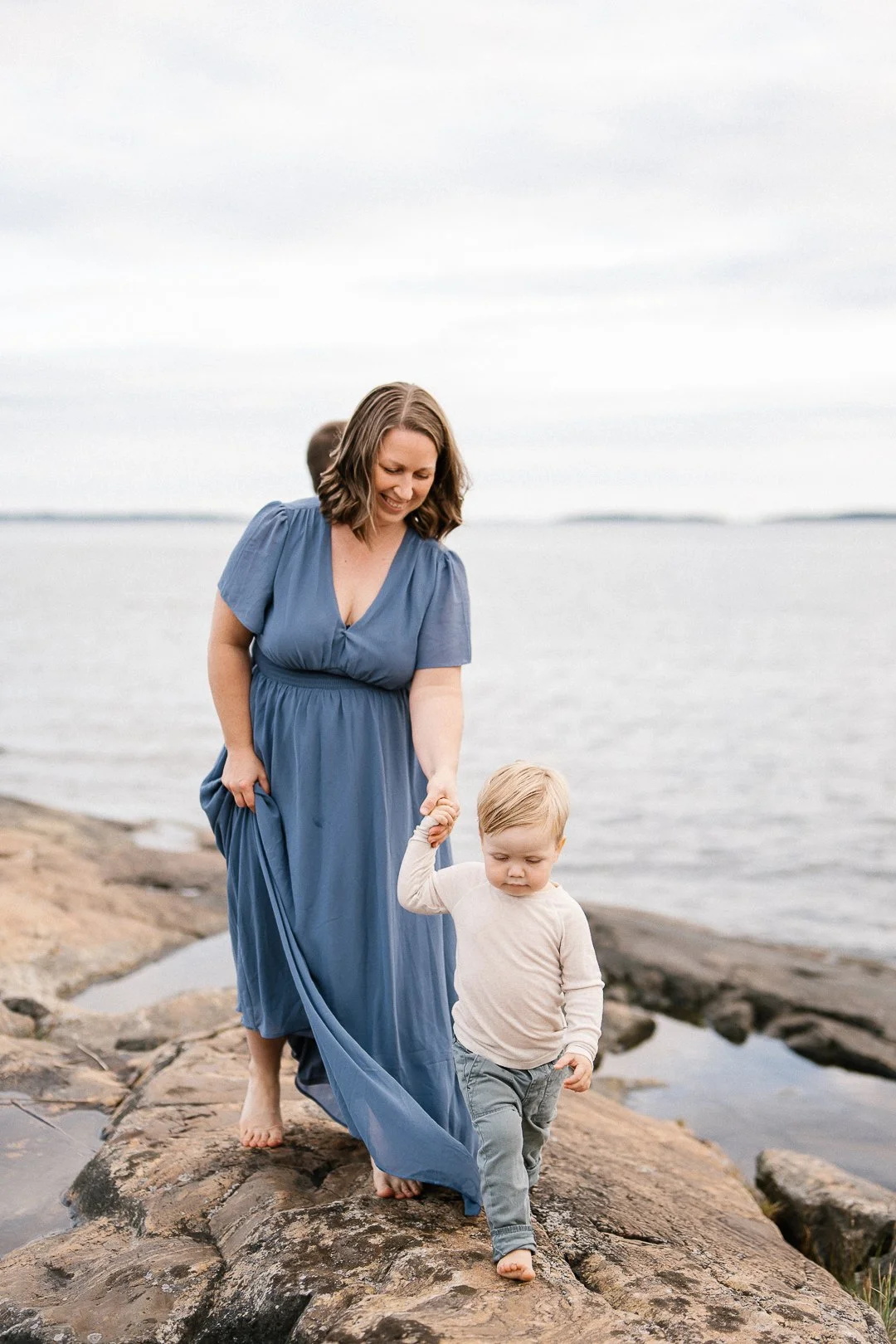 Nainen ja lapsi kävelevät rannalla. Nainen hymyilee ja pitää lapsen kättä. Heillä on vaatteet, naisen puku on sininen ja lapsen paita vaaleankeltainen.