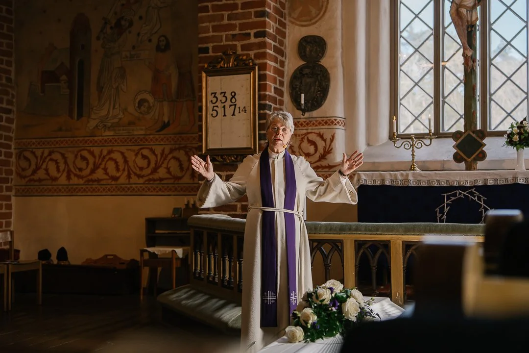 Pappi tai pastorin nainen, joka seisoo kirkossa ja tulkitsee tai rukoilee, pitäen kädet levossa. Taustalla ikkunasta näkyy luonnon valoa ja kirkon sisustusta, kuten kynttilöitä, kukkia ja kirkkovehkeitä.