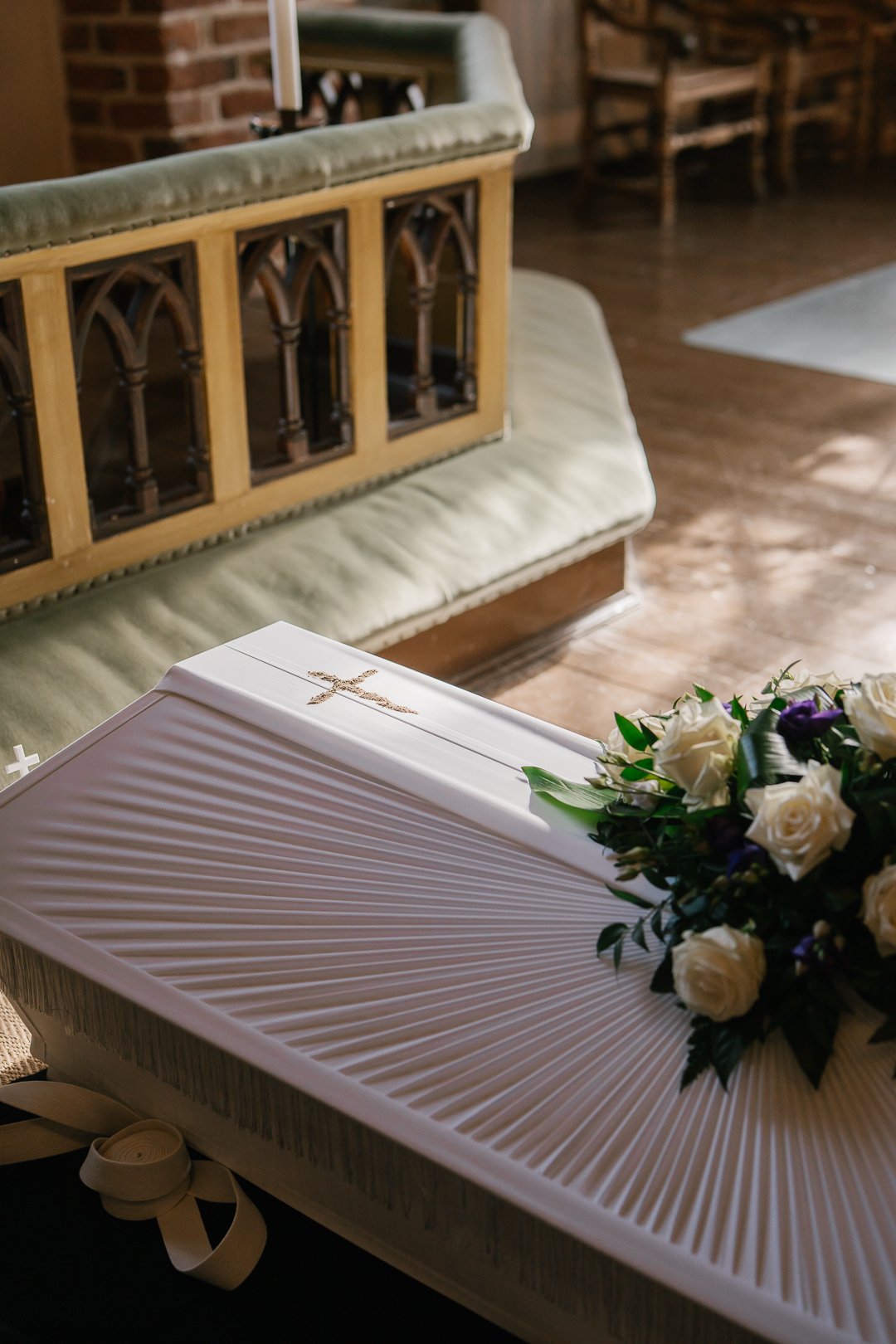 Ristimäinen valkoinen kirja ja kukkavalkoinen kimppu, jonka päällä on risti, sijaitsevat pöydällä, taustalla kiviseinä ja beigeä verhoilua.