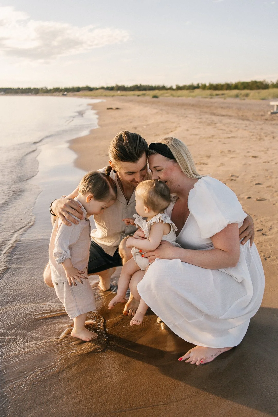 Perhe hymyilee ja katselee toisiaan rannalla, lapset ja aikuiset, auringonlaskun aikaan.