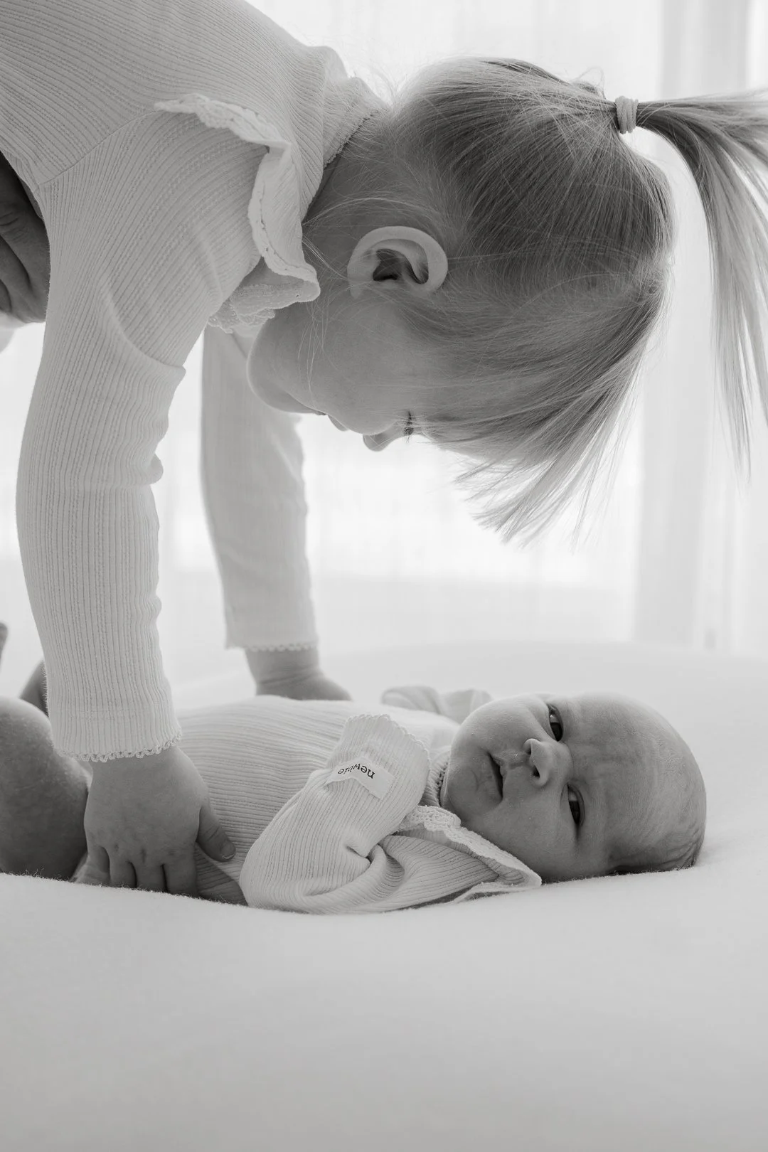 Vauva makaa alustalla ja hymyilee, kun vanhempi leikkii tai kommunikoi hänen kanssaan päänsä alas päin. Kuvassa lapset ja vanhempi ovat pukeutuneita vaaleisiin vaatteisiin ja tausta on valkoinen, valoisassa huoneessa, mustavalkoisena.