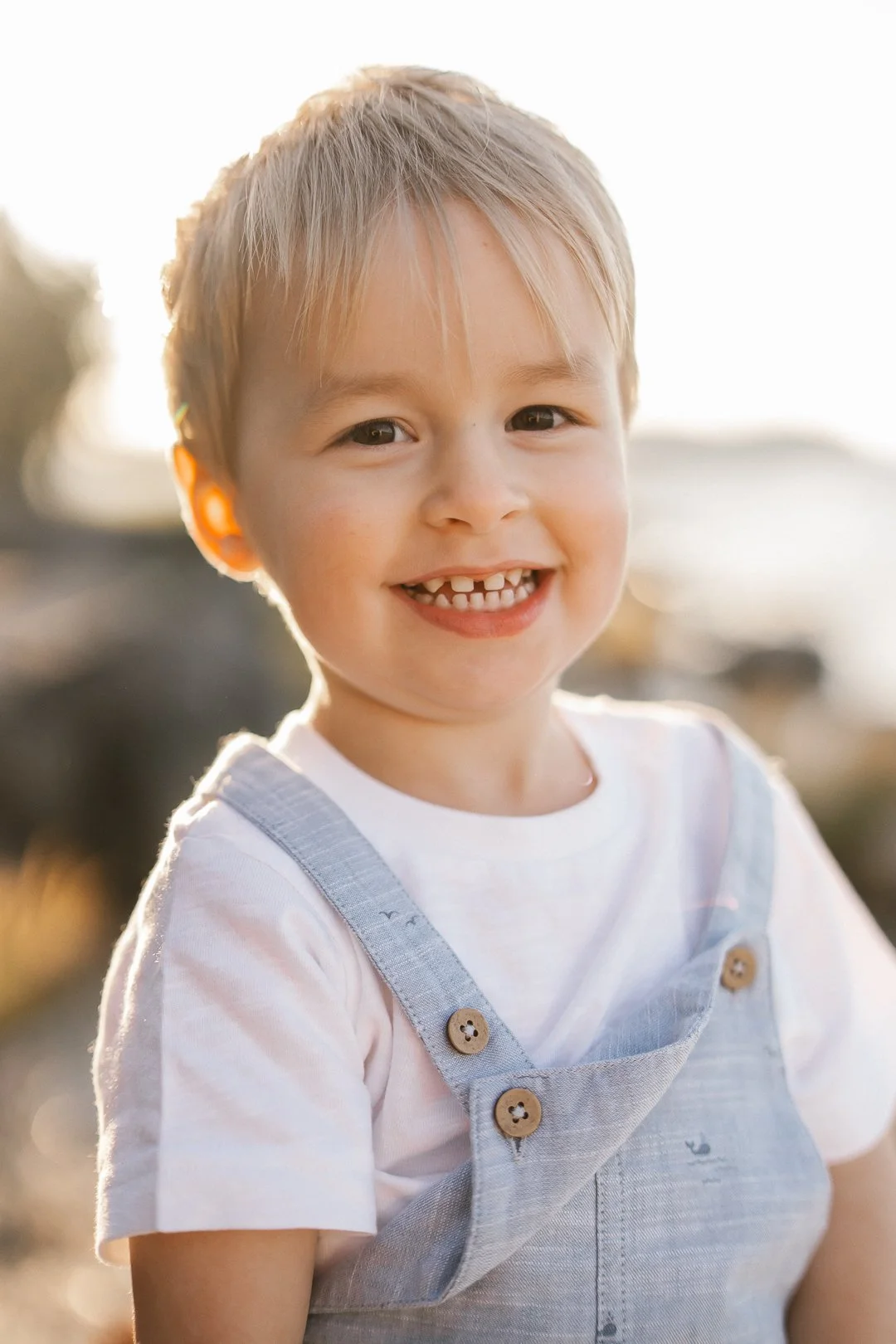 Hymyilevä pieni poika, jolla on vaaleat hiukset, pukeutuneena valkoiseen T-paitaan ja harmaisiin haalareihin, auringonpaisteessa ulkona.