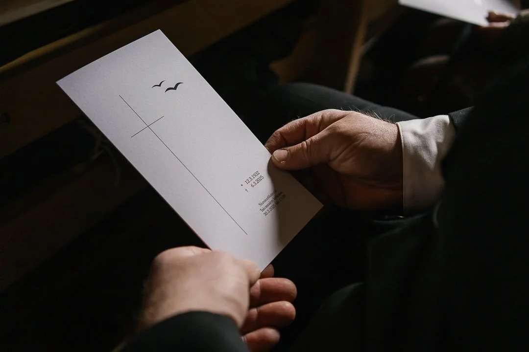 Käsivarsi ja käsi, joka pitää valkoista paperia, jossa on pieniä mustia lintu- ja ristimuotoisia piirroksia sekä tekstiä ja numeroita. Tausta on pimeä, istuimen tai penkin selkänoja näkyy taustalla.