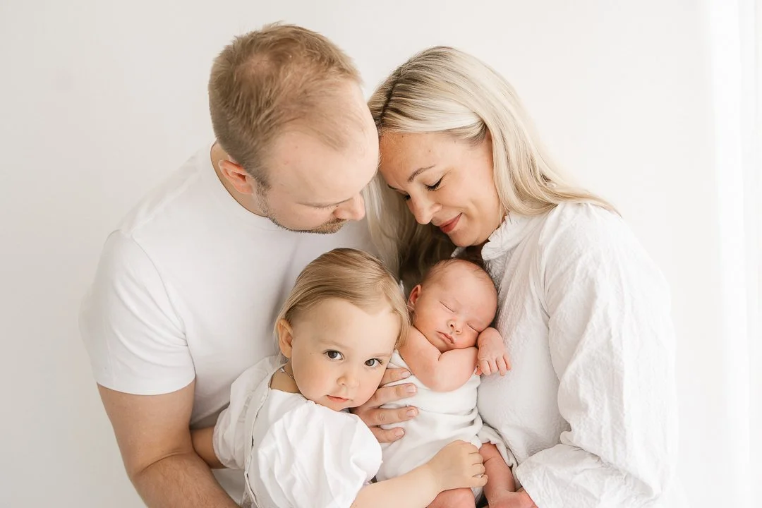 Perhe, isä ja äiti iloitsivat uudesteruksesta, kaksi pientä lasta, isompi tyttö ja pieni vauva, pitelevät vauvaa, taustalla vaalea seinä.