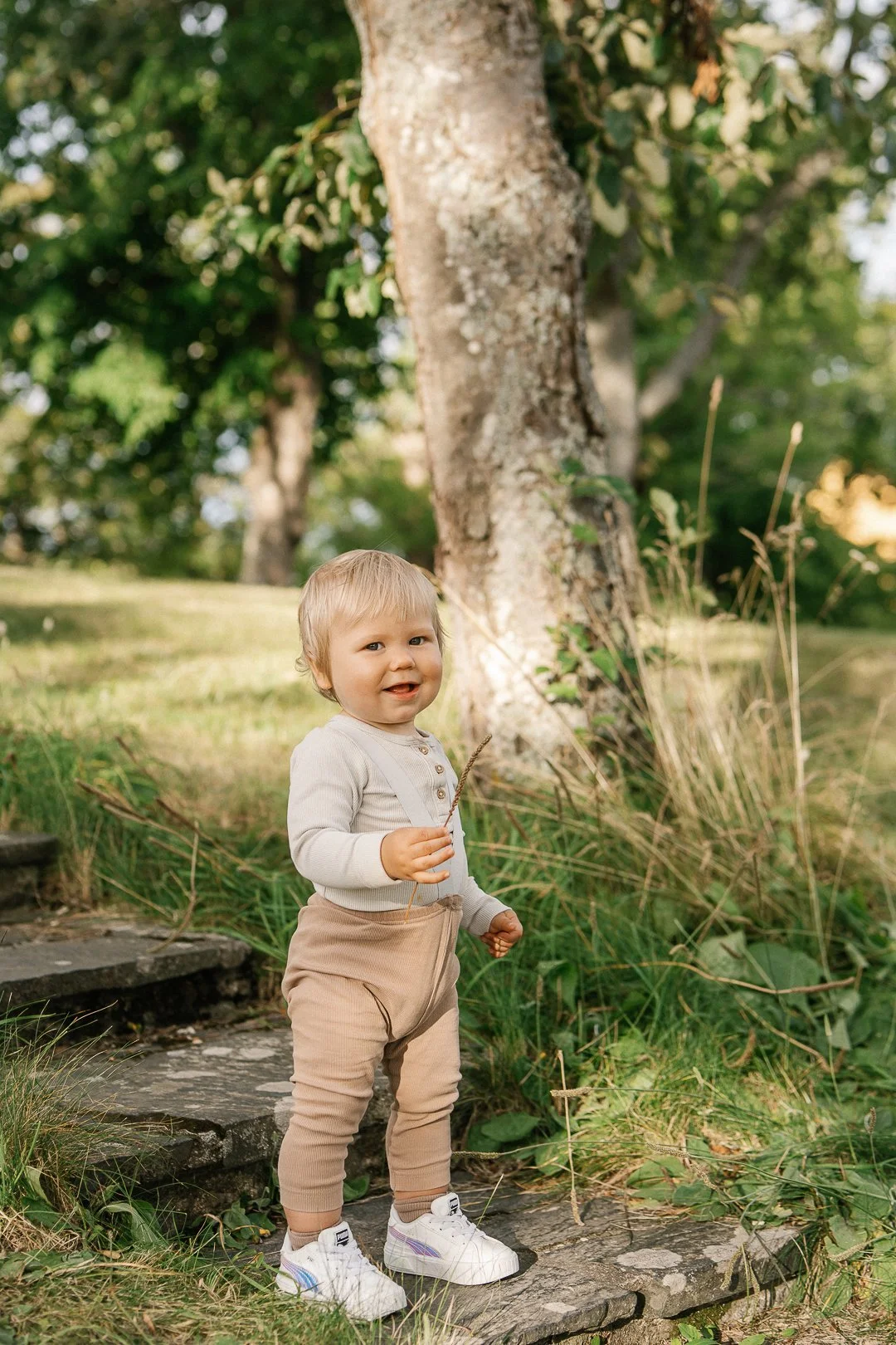 Vauva seisoo pihalla kivetyksellä, taustalla puita ja vihreitä kasveja.