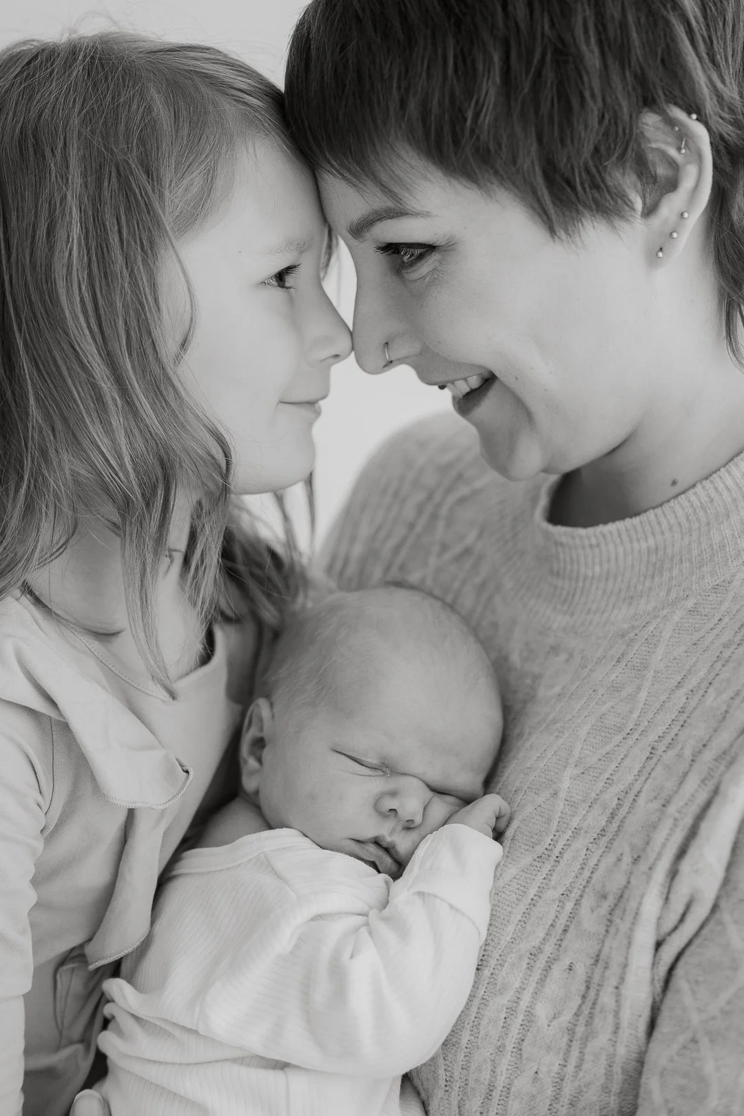 Kuvassa on perhe, jossa pieni vauva nukkuu äidin ja isän välillä. Äidin ja isän otsat ovat kosketuksissa, ja he katsovat toisiaan hymyillen.