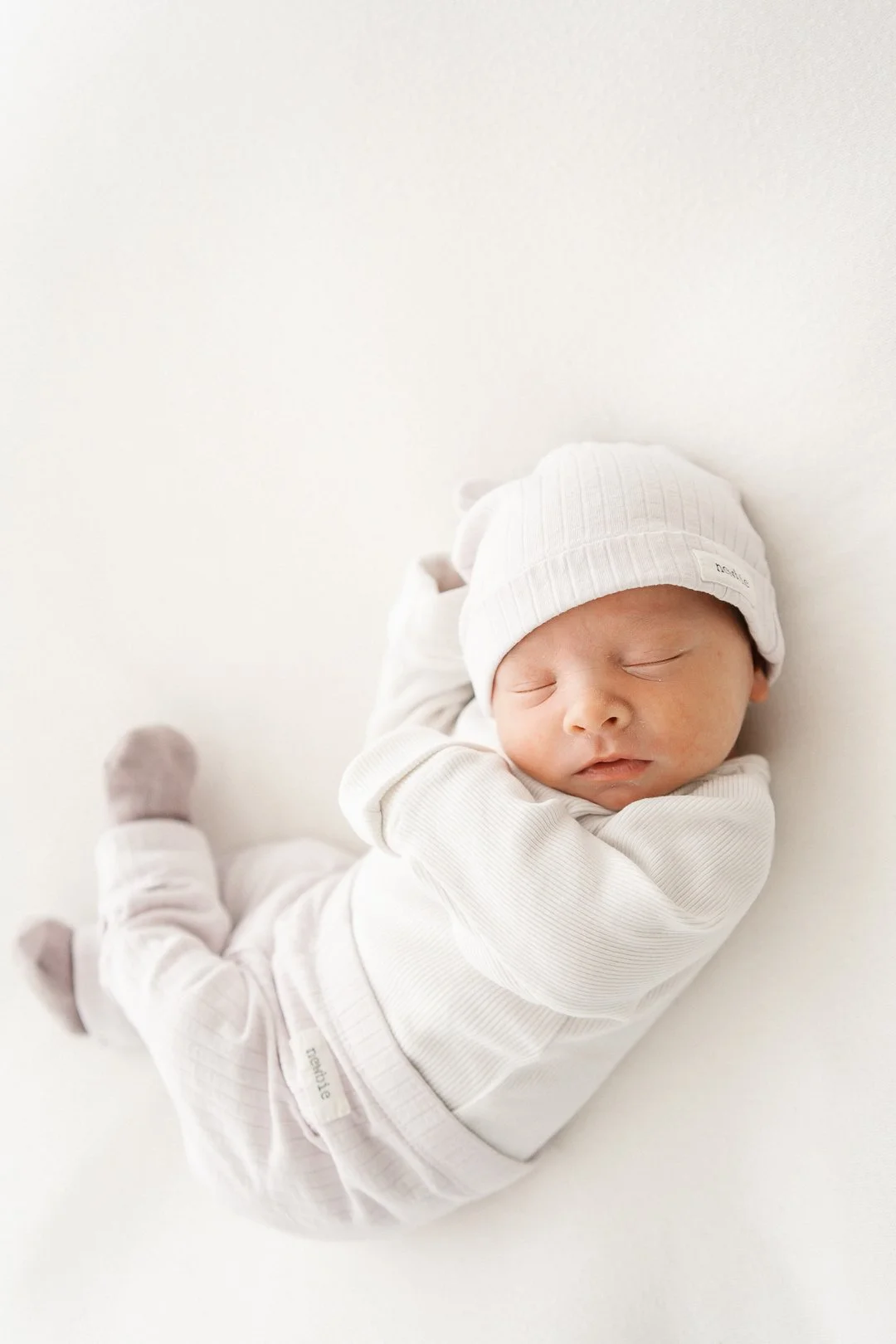 Vauva nukkuu, pukeutunut valkoisiin vaatteisiin ja valkoiseen pipoonsa, joka on leikattu nauhamaiseksi.