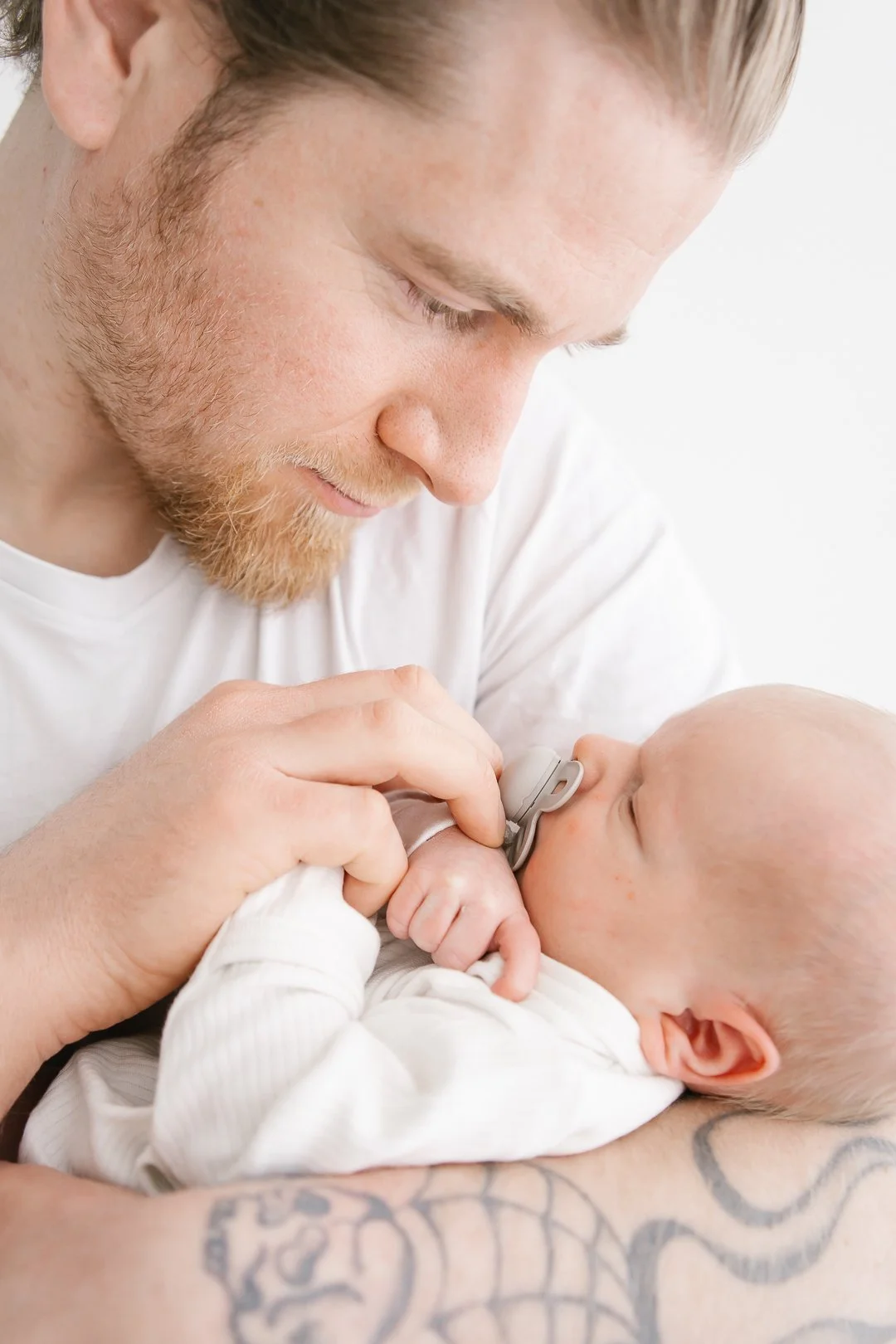 A man with a beard and tattoos holding a baby with a pacifier in its mouth, close-up shot.