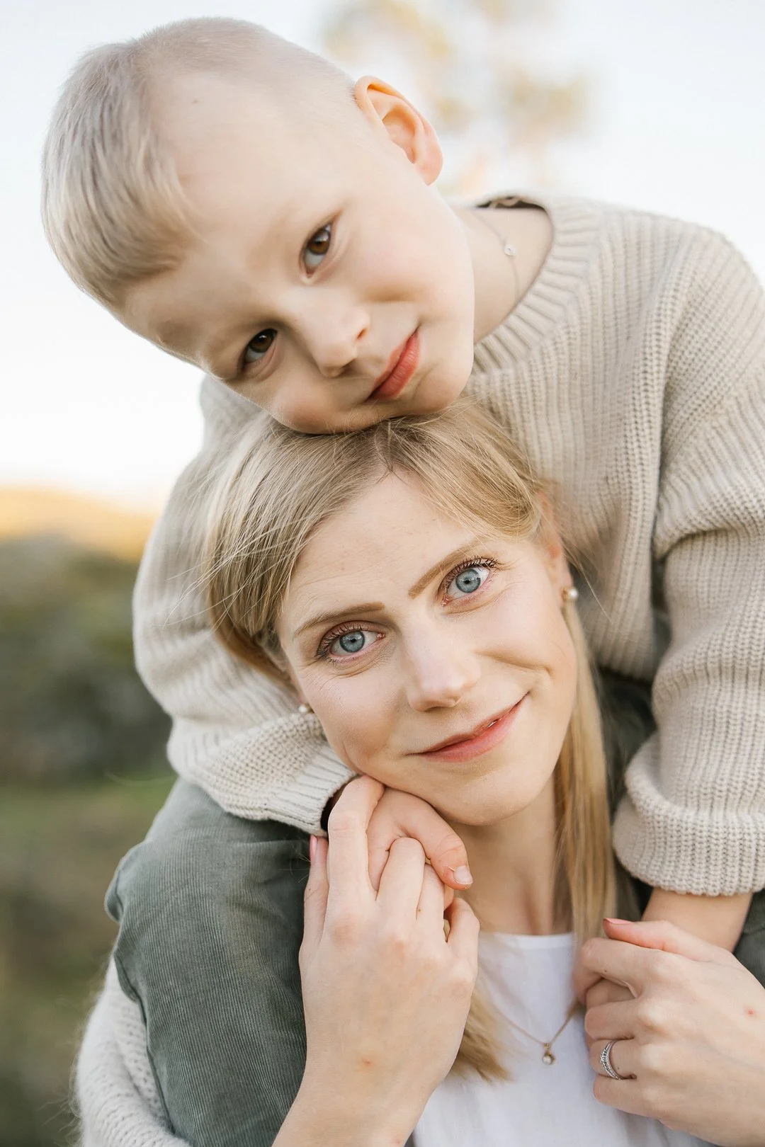 Äiti ja lapsi ulkona, lapsi istuu äidin selän päällä, katsoo suoraan kameraan, äiti hymyilee ja pitää päätä kädessään, taustalla luonnonäkymä.