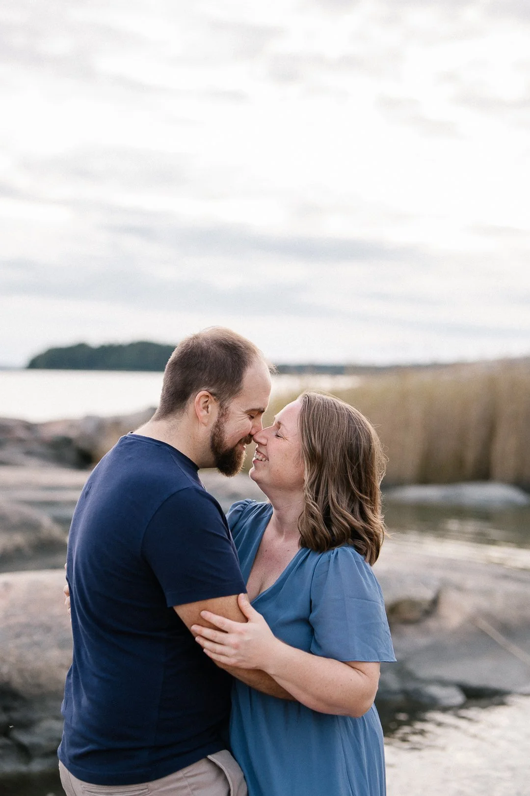 Pariskunta hymyilee ja nojaa otsat eteen rannalla, taustalla meri ja pilvinen taivas.