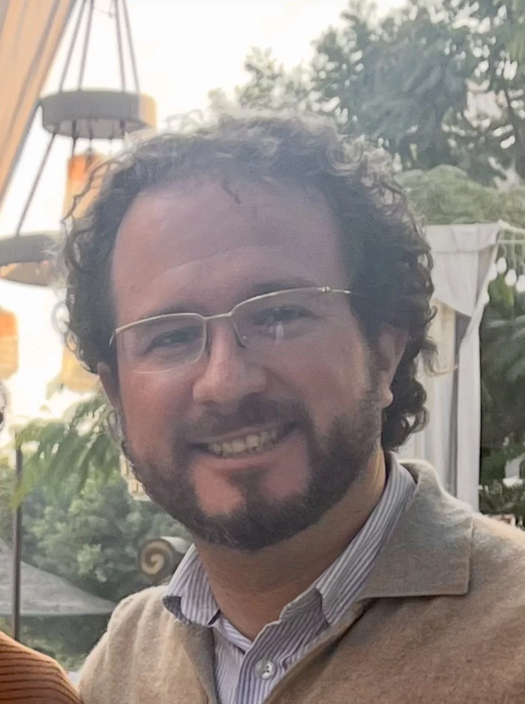 Ein lächelnder Mann mit Brille und braunem Bart, trägt ein cremefarbenes Pullover und ein gestreiftes Hemd, im Hintergrund Pflanzen und eine Terrasse.