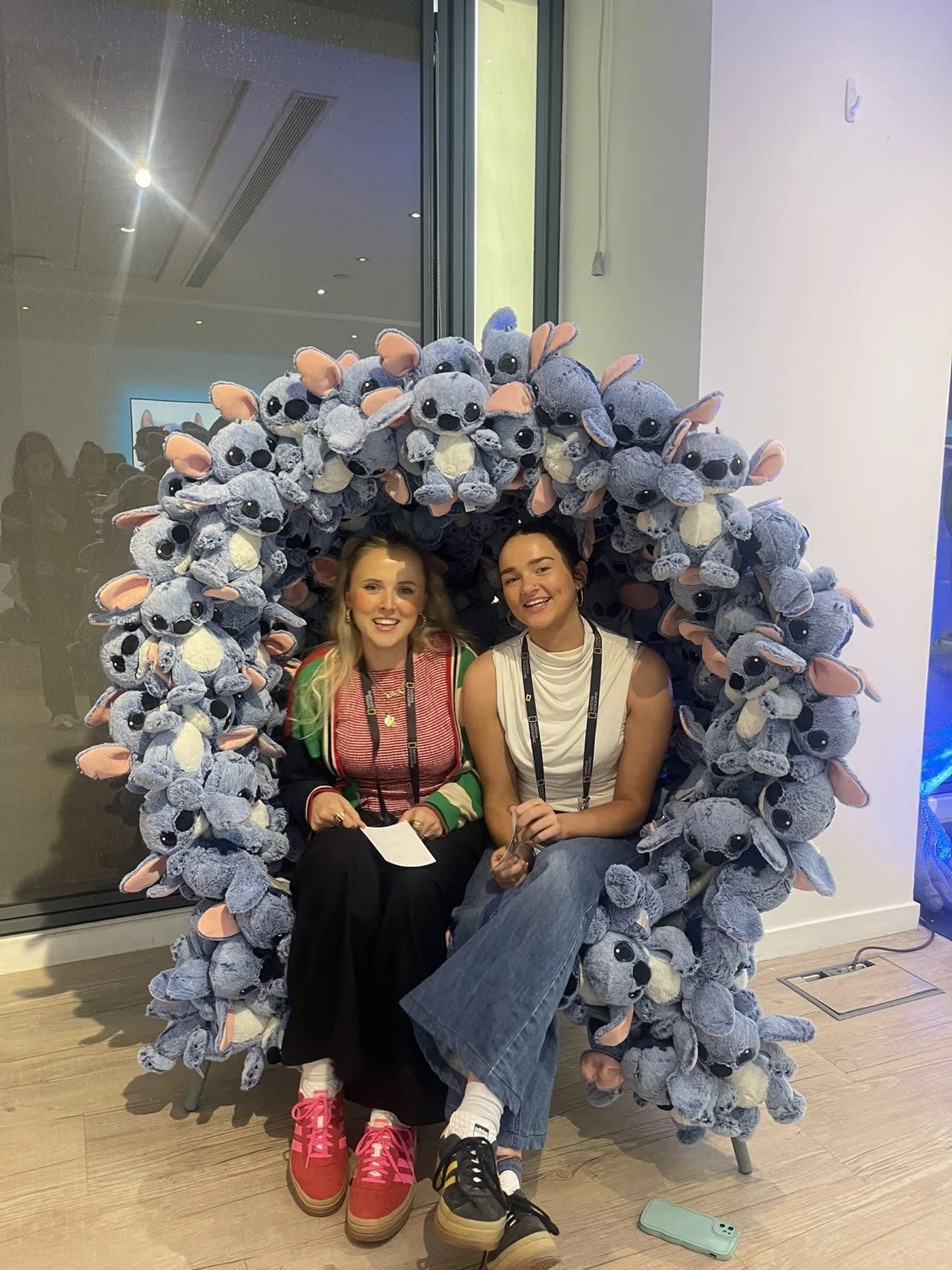 Two women sitting inside a large chair made of plush toy stitch dolls, smiling at the camera. One woman has long blonde hair and is wearing a colorful striped sweater and red shoes. The other has dark hair and is wearing a sleeveless top, jeans, and white shoes. They are in a modern indoor space with a glass window behind them and people reflected in the glass.