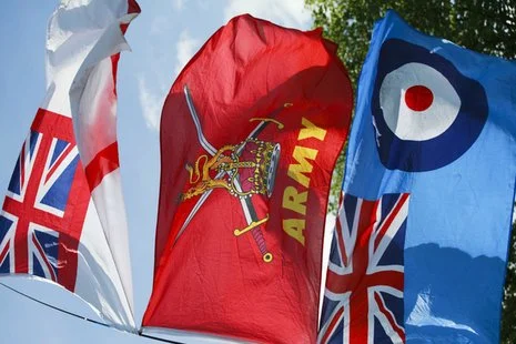 Multiple flags, including the British Union Jack, a red flag with a knight's helmet and the word 'ARMY', and a blue flag with a target design, waving in the wind outdoors.