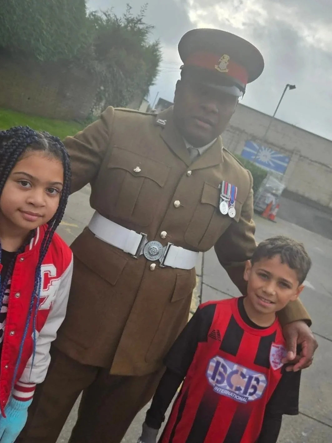 A man in military uniform with medals, standing outdoors with two children, a girl and a boy, both wearing sports jerseys.