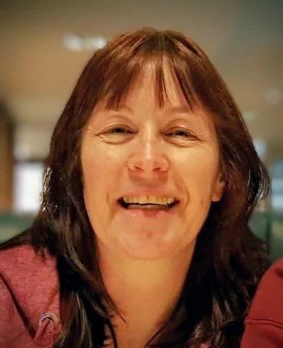 Close-up of a smiling woman with shoulder-length brown hair and bangs, wearing a maroon top, in an indoor setting.