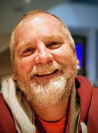 A smiling man with a beard and mustache, wearing a red hoodie with a gray shirt underneath, in an indoor setting.