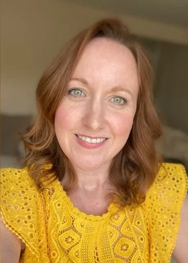 A woman with shoulder-length wavy red hair and blue eyes smiling at the camera, wearing a bright yellow, lace-patterned top.
