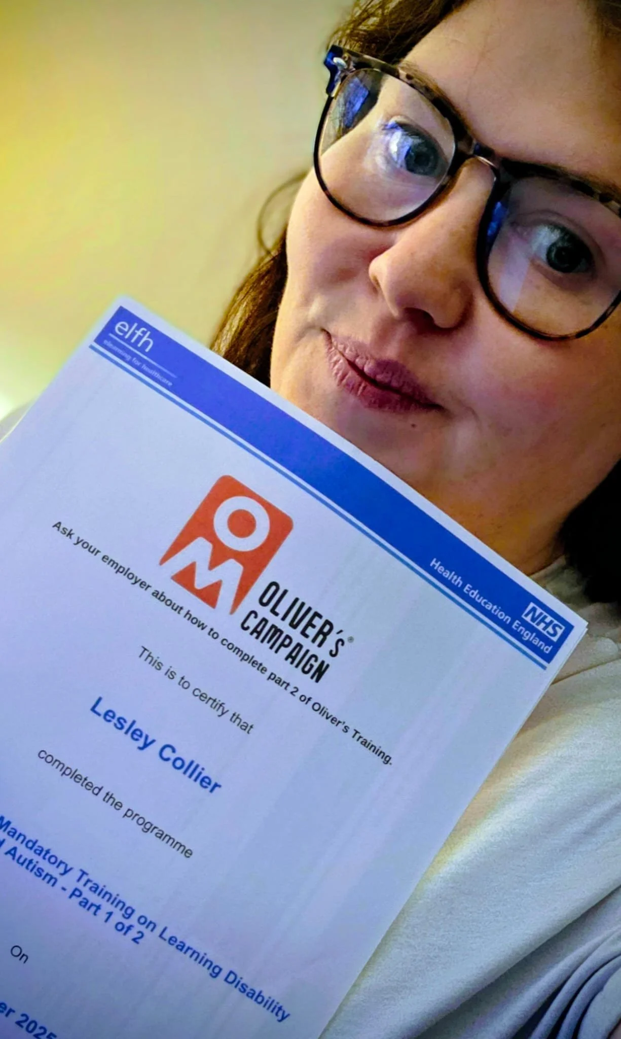 A woman wearing glasses holds a certificate from NHS England's Oliver's Campaign, verifying her completion of part 2 of Oliver's Training on learning disability and autism.