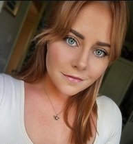 Close-up of a woman with red hair, blue eyes, wearing a white top and a necklace with a small animal pendant, smiling at the camera.