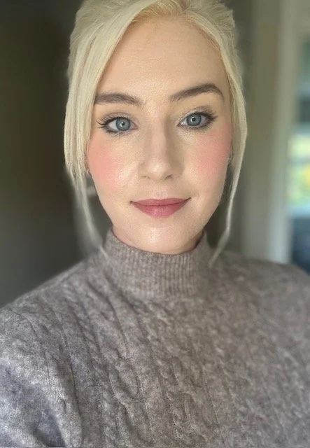 A woman with blonde hair, blue eyes, and fair skin, wearing a gray knit turtleneck sweater, smiling inside a room with natural light.