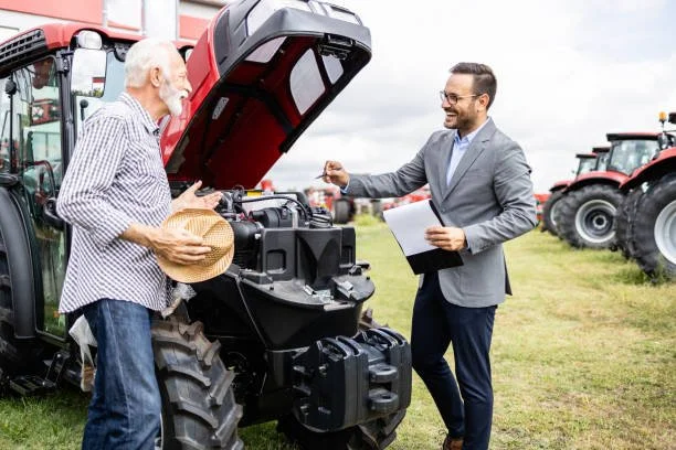 An older man with a hat under his arm talking to a younger man in a suit holding a clipboard, standing next to a tractor with its hood open on a farm with other tractors in the background.
