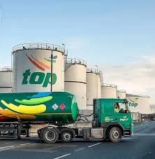 A green tanker truck with a banana graphic and the word 'top' on the side, driving on a highway near large industrial storage tanks.