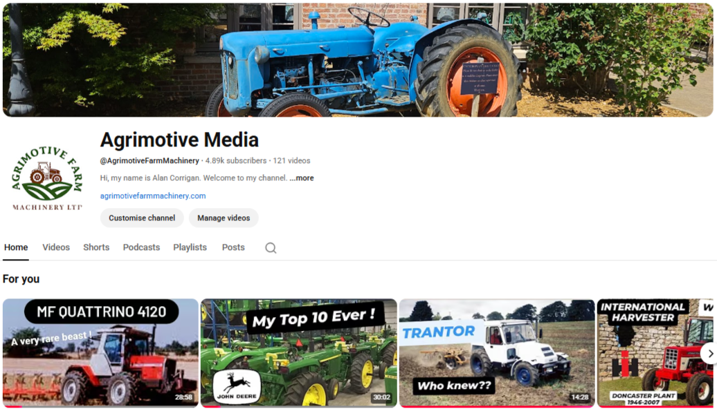 A vintage blue tractor on display outdoors with trees and a brick building in the background.