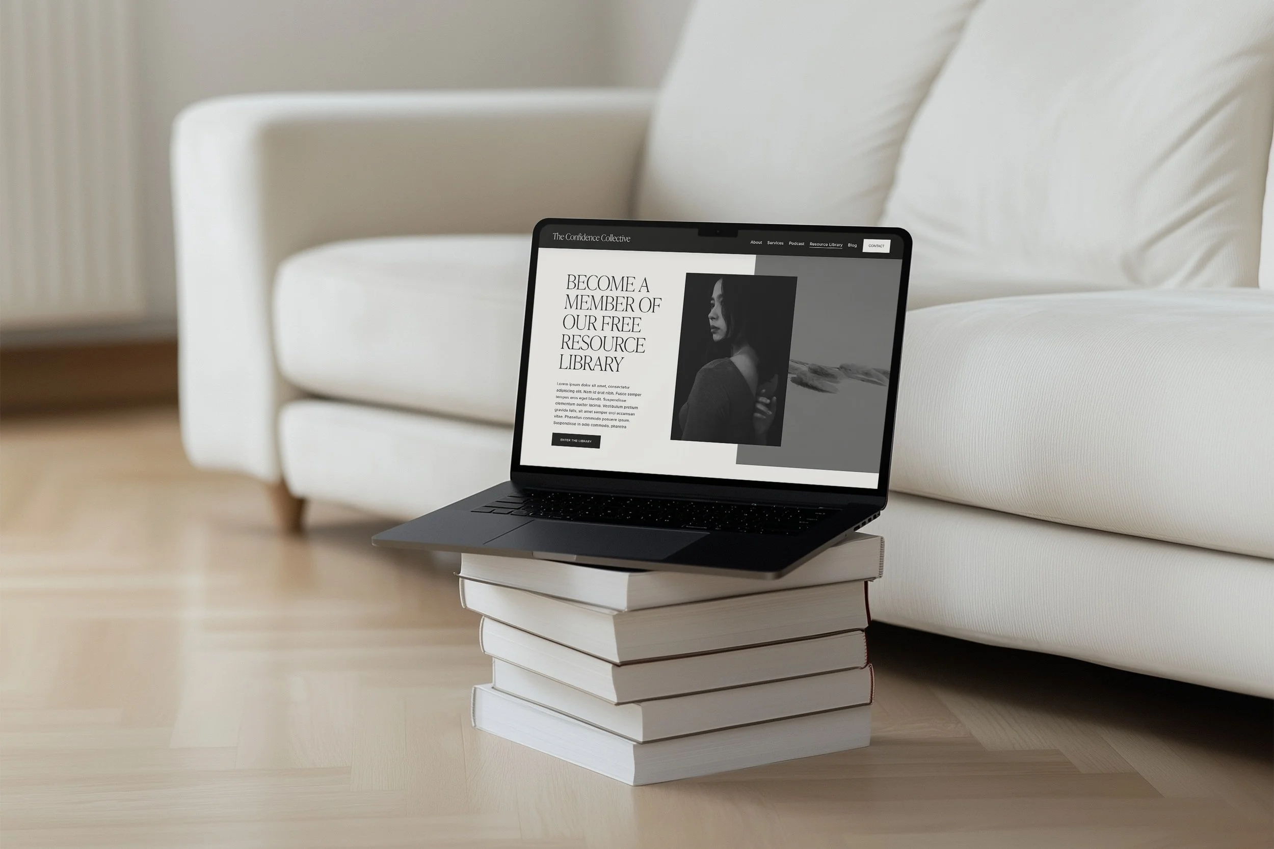 A laptop placed on top of a stack of six white books on a light wood floor, in front of a white sofa. The laptop screen displays a website with the text 'Become a Member of Our Free Resource Library' and a black and white photo of a woman.