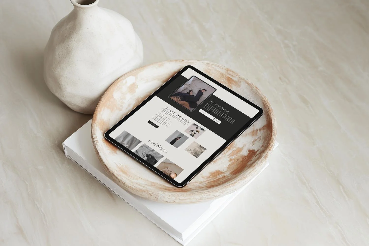 A tablet on top of a closed book inside a ceramic bowl, with a decorative vessel in the background.
