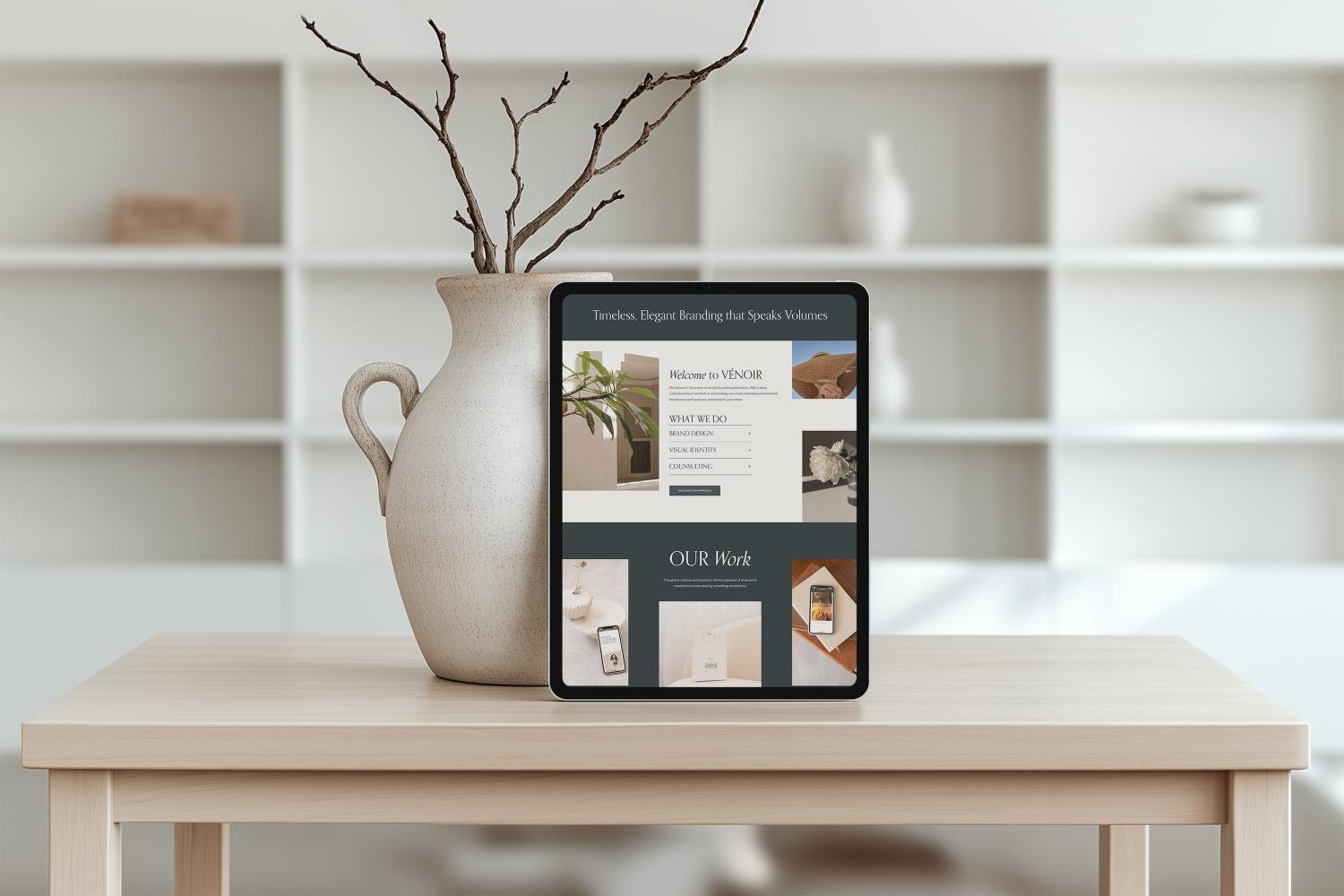 A tablet displaying a website, placed on a wooden table beside a large beige vase with leafless branches, in a minimalist room with white shelving in the background.